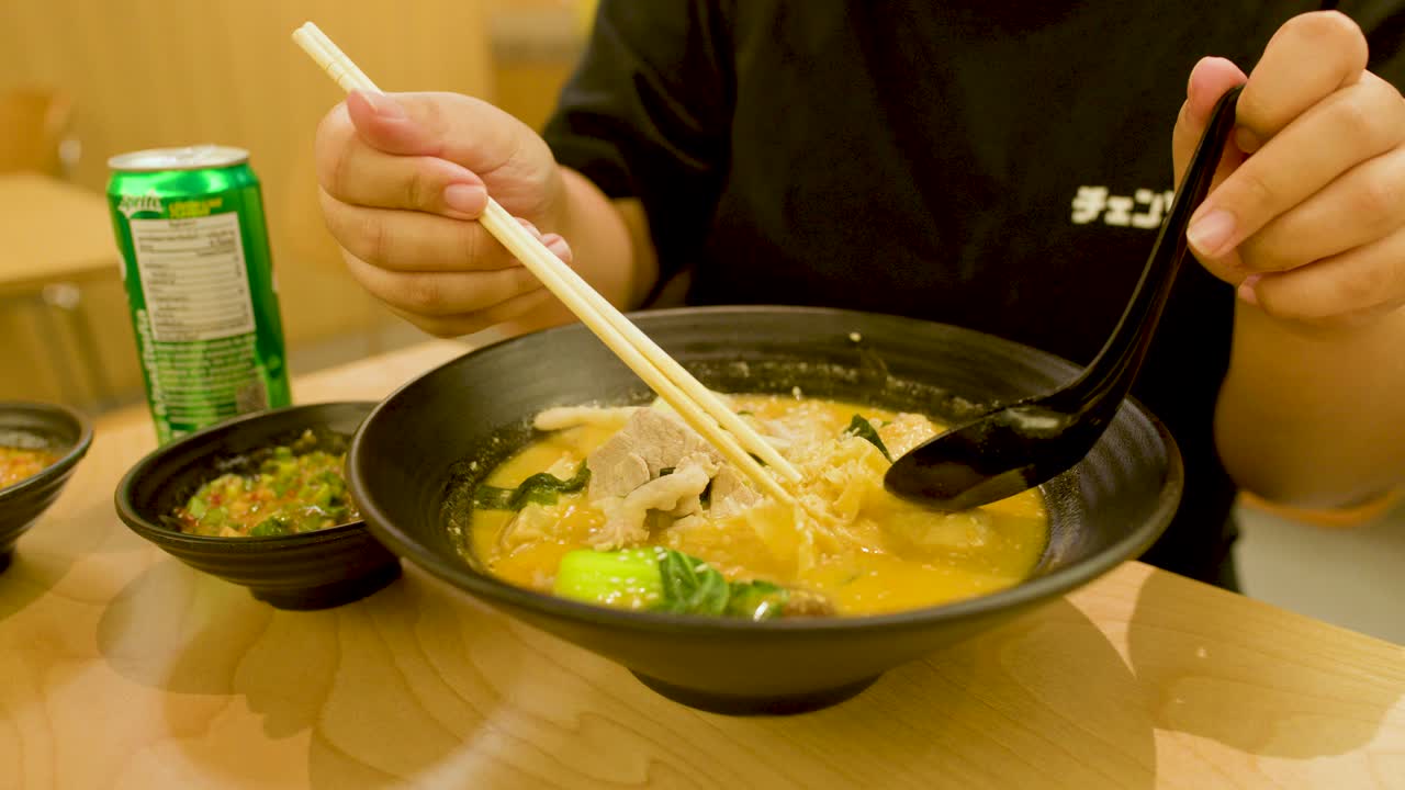 Person uses chopsticks and spoon to eat hot noodle soup in brightly lit casual restaurant
