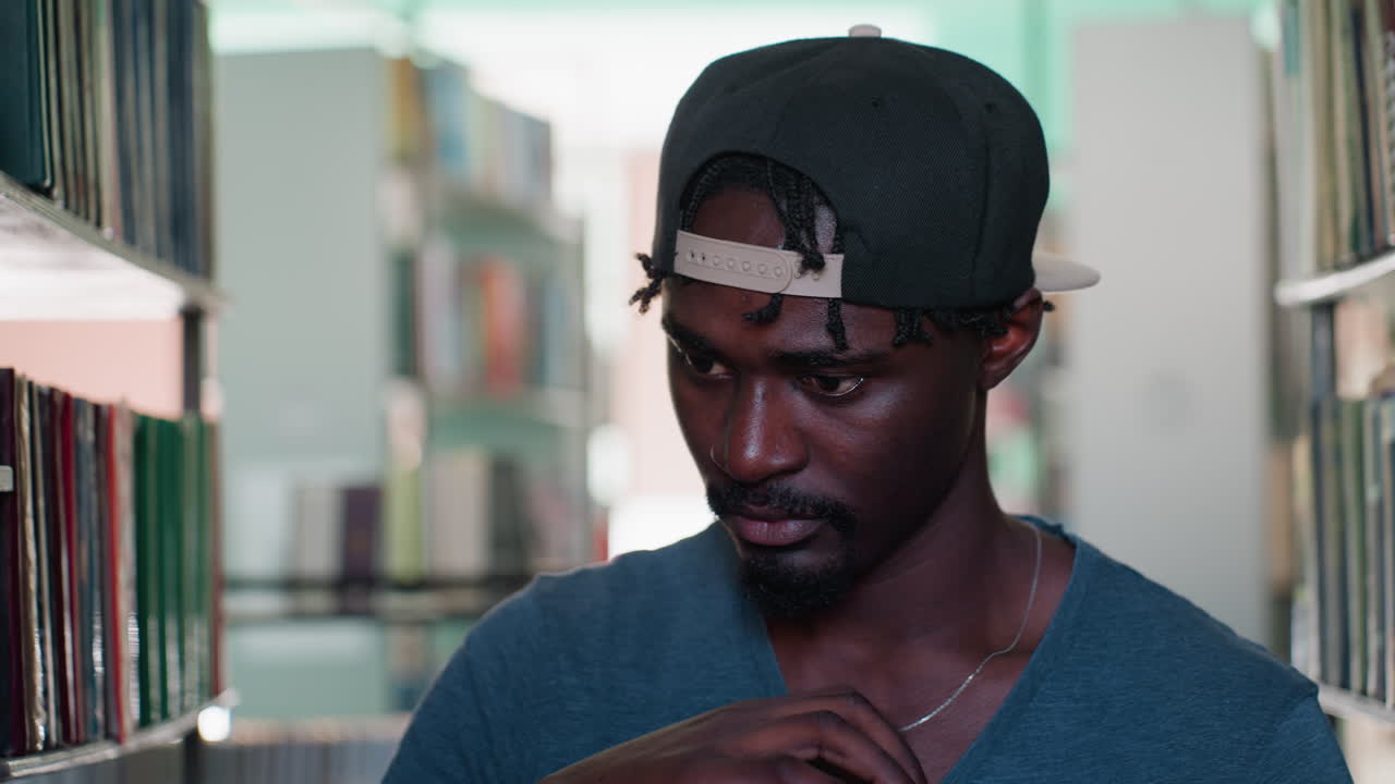 Close up of young student wearing cap backwards, holding stack of books in library, gently tapping cover of top book while looking to the side with thoughtful expression among blurred bookshelves