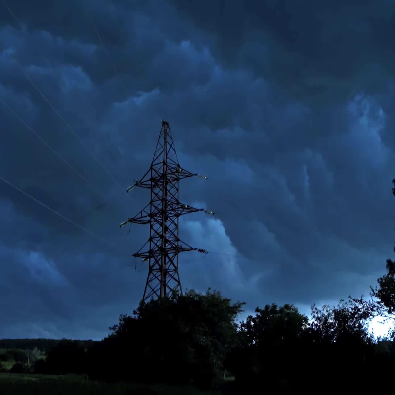 Stormy weather in nature. Dark clouds moving on the sky over the tower of high voltage electricity. Strong wind shaking trees in the evening.