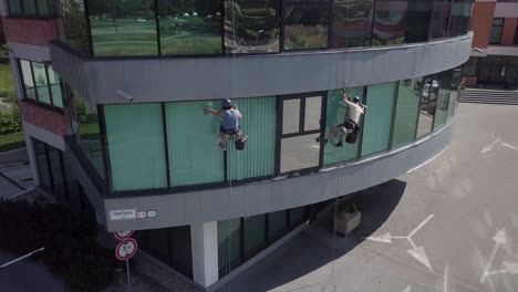 Rotación-Aérea-Lenta-Alrededor-De-Los-Trabajadores-Escaladores-Industriales-Que-Limpian-Las-Ventanas-De-La-Oficina