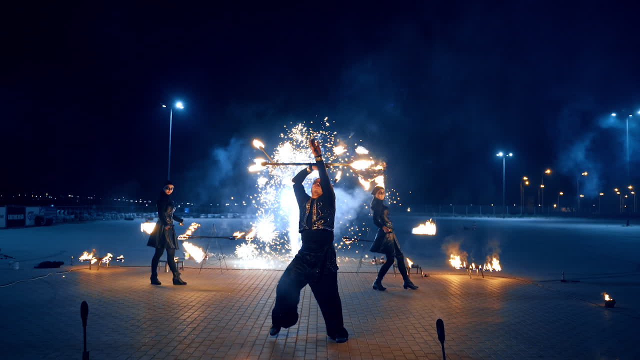 Man in black costume dances rotating the burning stick. Two female dancers turn the props in fire at backdrop.