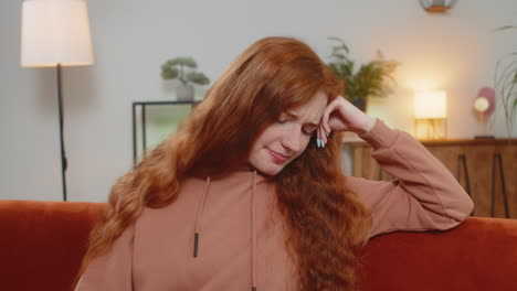 upset lonely young woman with long red hair thinking about problems sitting on sofa in living room