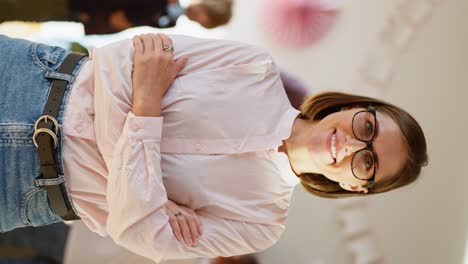 Vertical-video-portrait-of-a-happy-female-teacher-with-a-bob-hairstyle-in-glasses-with-blue-eyes-in-a-pink-shirt-who-smiles-with-her-arms-crossed-on-her-chest-and-looks-at-the-camera-against-the-background-of-a-club-for-preparing-children-for-school