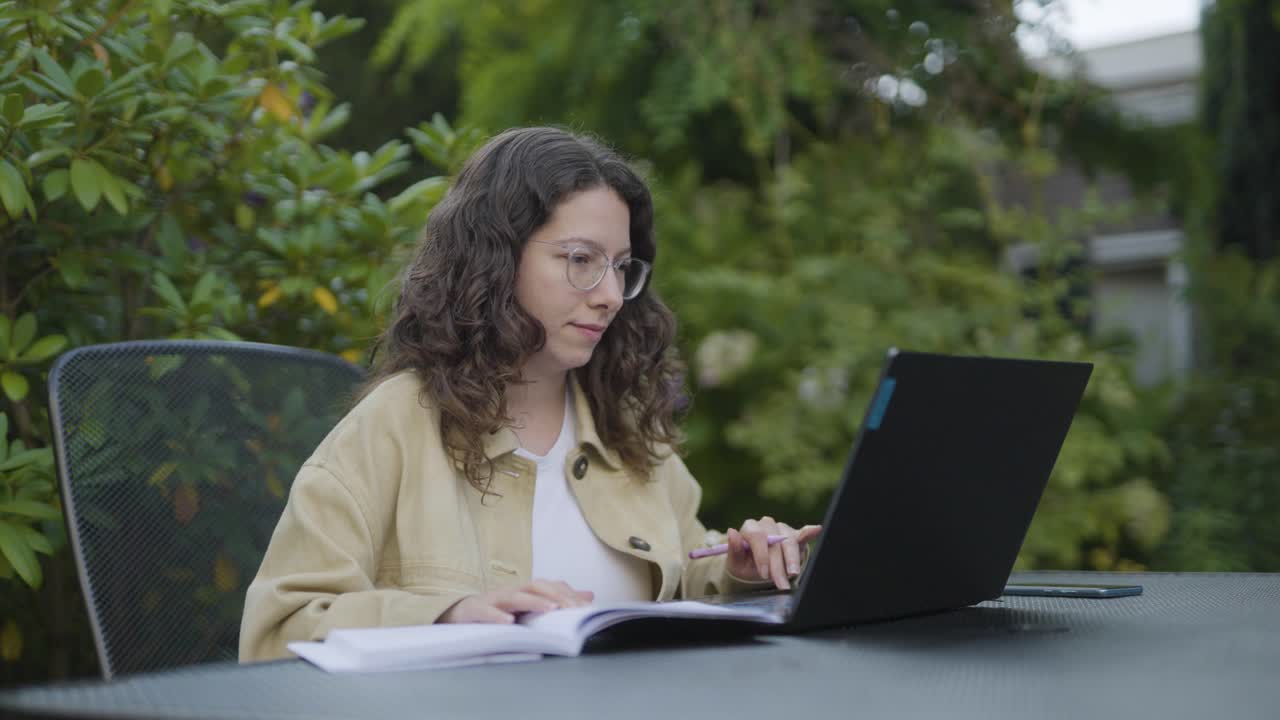 Focused young woman with glasses studying outside at a garden table, using a laptop and book in a natural green environment