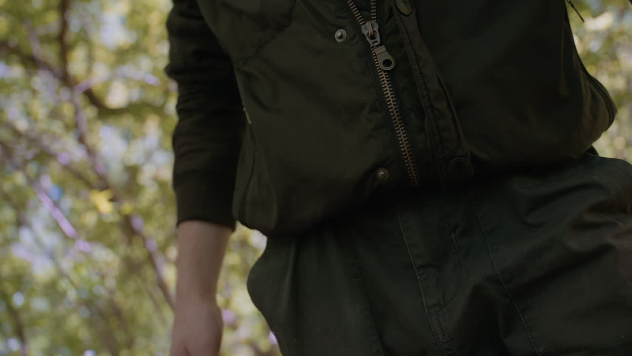 Close-up view of adventurer zipping jacket halfway and folding the tip while dragging it down to cover the belt, with sunlight filtering through trees, preparing for the next stage of his journey