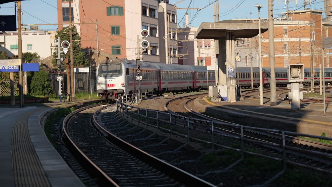 Slow motion clip of an Italian train approaching Civitavecchia station on a curved track, with warm sunlight highlighting urban surroundings and railway infrastructure