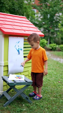 little boy painting with his finger on the sheet of paper stuck to the wall of playhouse. lovely child doing art hobby outdoors in summer. vertical video.