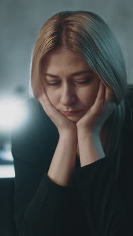 sad blonde lady leans on palms suffering from hard burnout at workplace with computer in semi-dark office in evening extreme close view slow motion