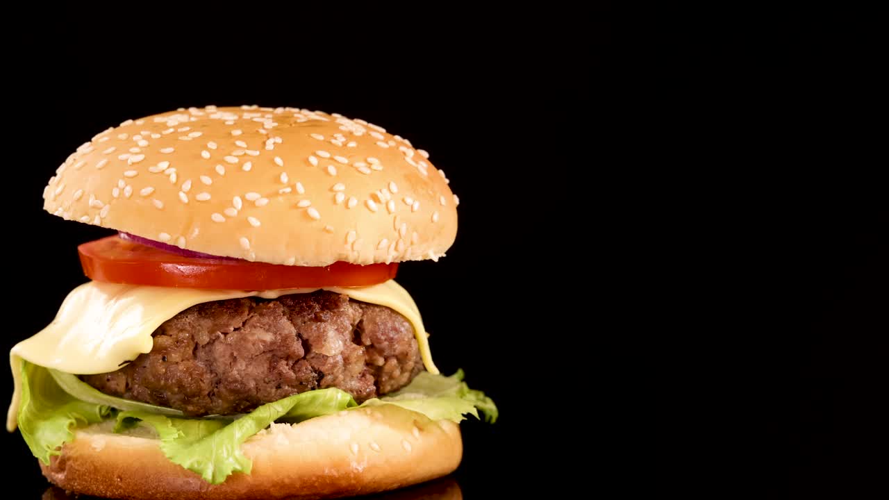 A classic beef burger with cheese, lettuce, and tomato rotates smoothly against a black background, illuminated by bright, even studio lighting