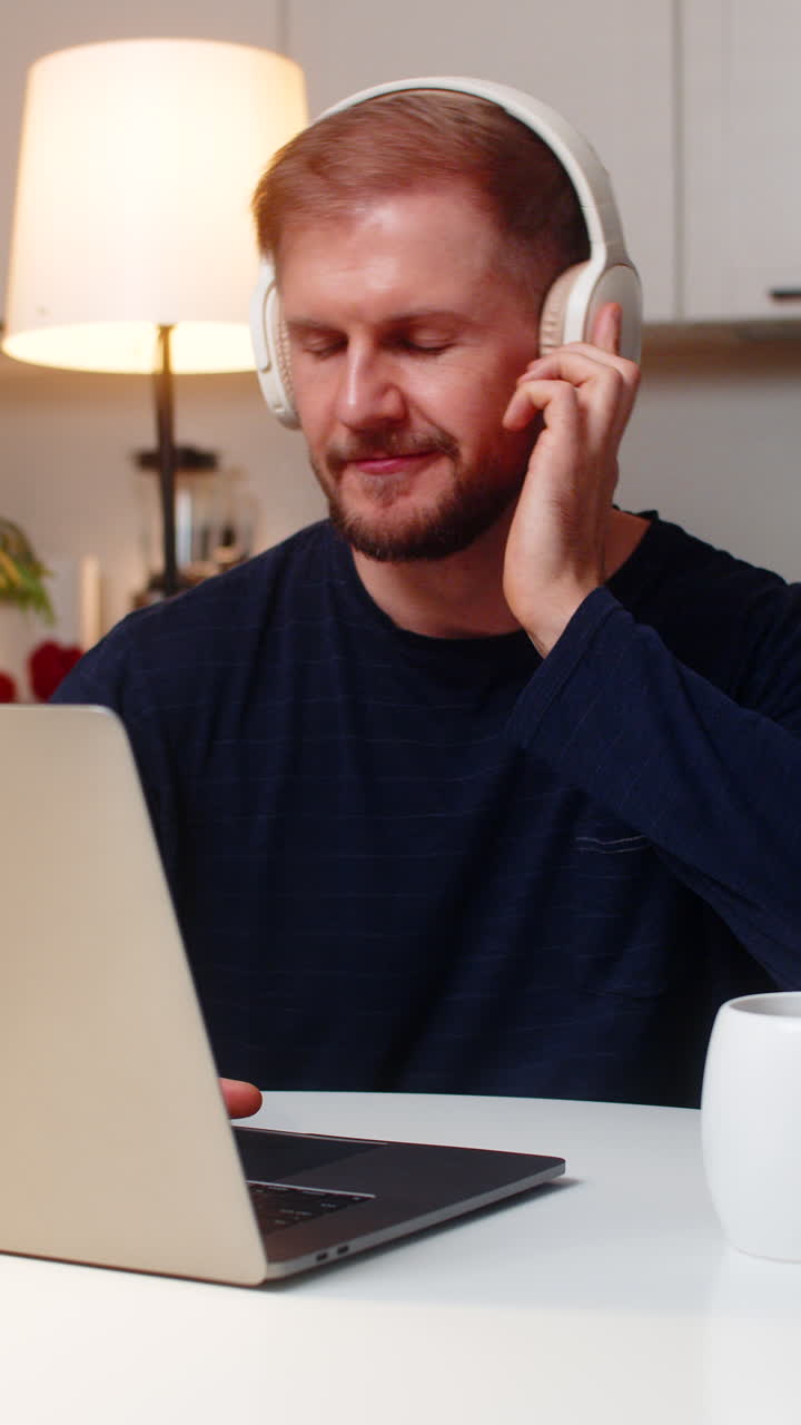 Ung mandlig freelancer med fokus på bærbar computer, der har trådløse hovedtelefoner på, drikker te og arbejder om natten indendørs
