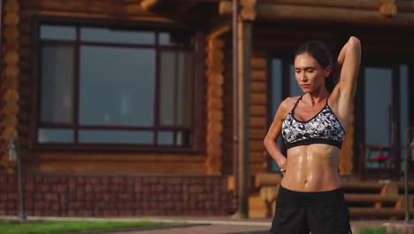 una mujer delgada y hermosa con ropa deportiva con una prensa abierta se está preparando para comenzar a entrenar en el césped cerca de su casa estirando sus brazos y hombros con ejercicios de calentamiento