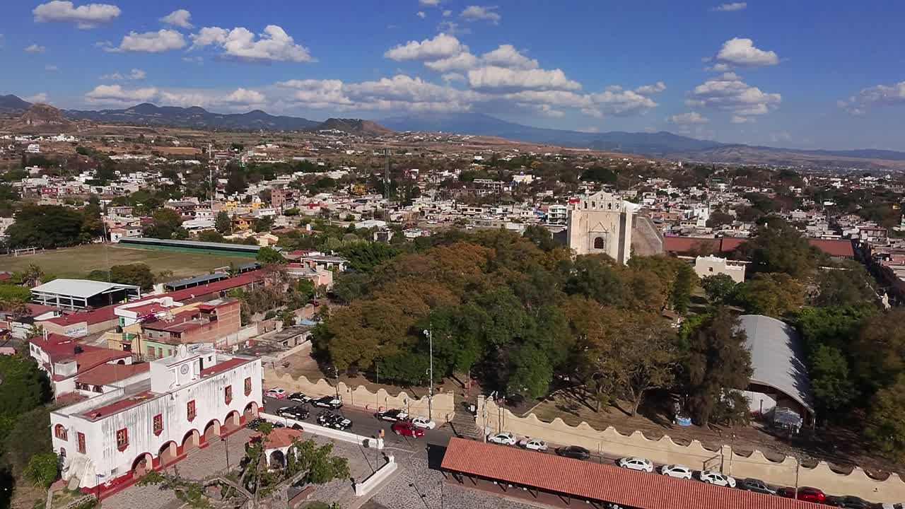 Aerial perspective of Tlayacapan, Morelos, captures the town's colonial architecture, church, vibrant marketplace, lush vegetation, and surrounding mountains under a clear blue sky