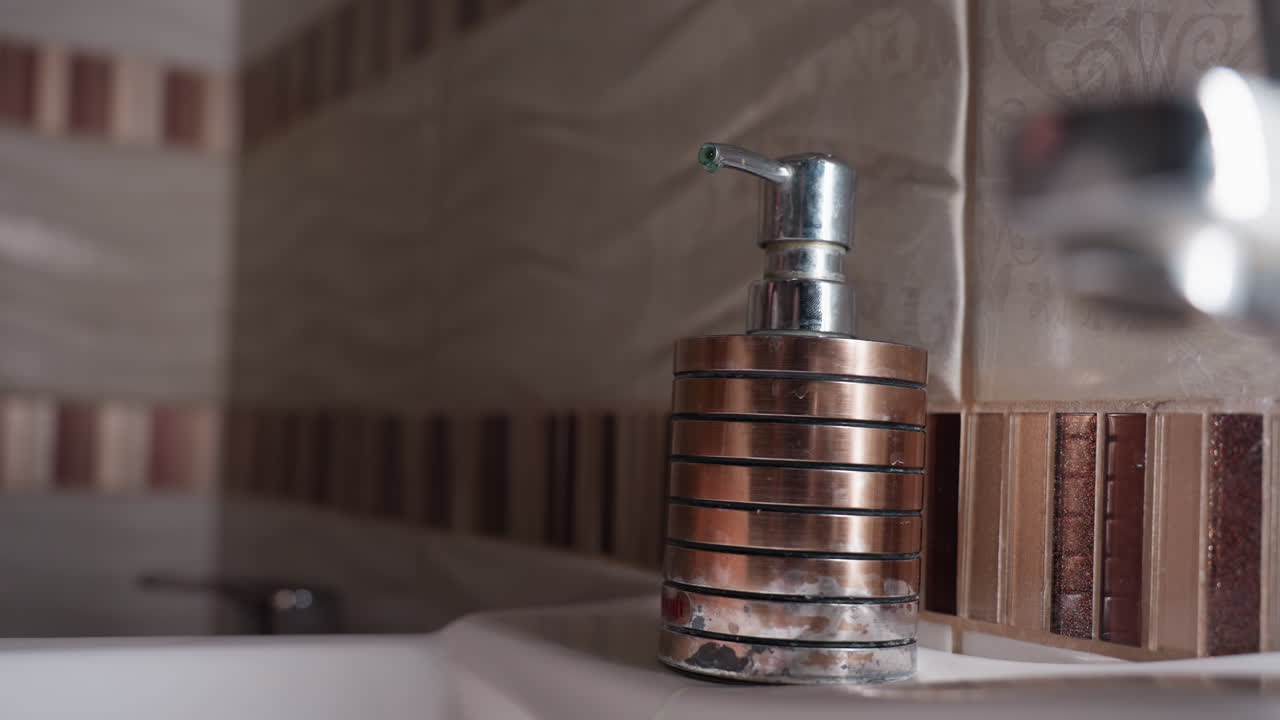 Hand view teenage hands at soap dispenser, one palm ready under tip while other presses pump, chrome bottle on sink beside tiled wall, hygiene action, clean routine, bathroom cleanliness concept