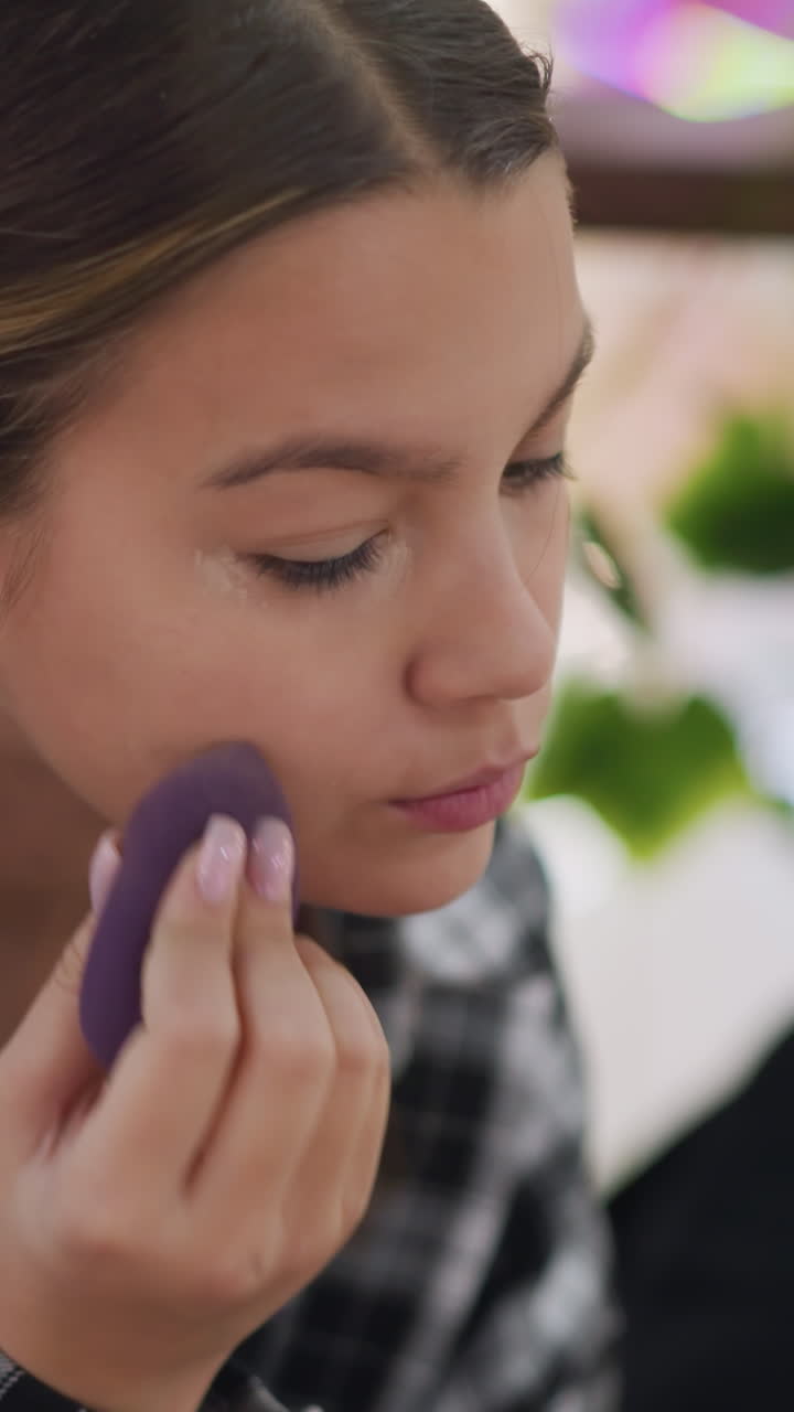 adolescente aplicando corrector con una esponja morada en la nariz, iluminación de backstage y retoque rápido, movimientos tranquilos y concentrados, manicura visible, primer plano, preparación para sesión de fotos o evento, cobertura sutil