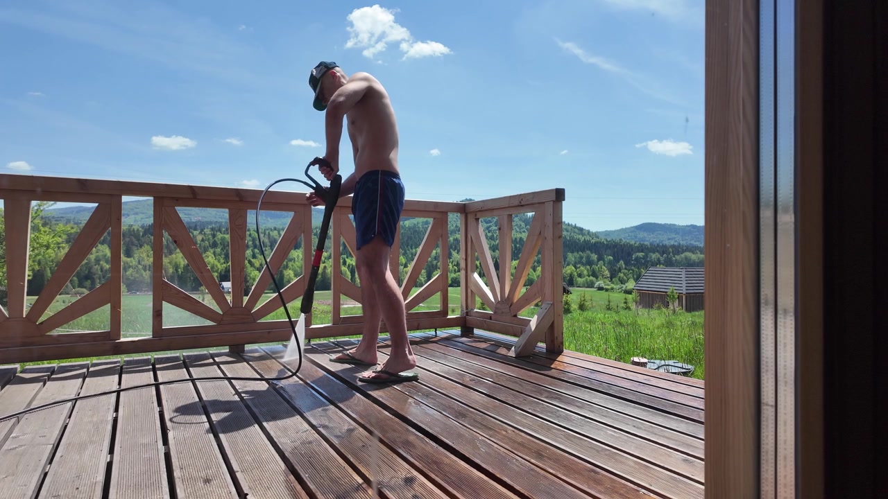 Uomo a torso nudo in pantaloncini blu pulisce una terrazza in legno con un'idropulitrice, con erba verde, foresta e cime montuose visibili sullo sfondo.