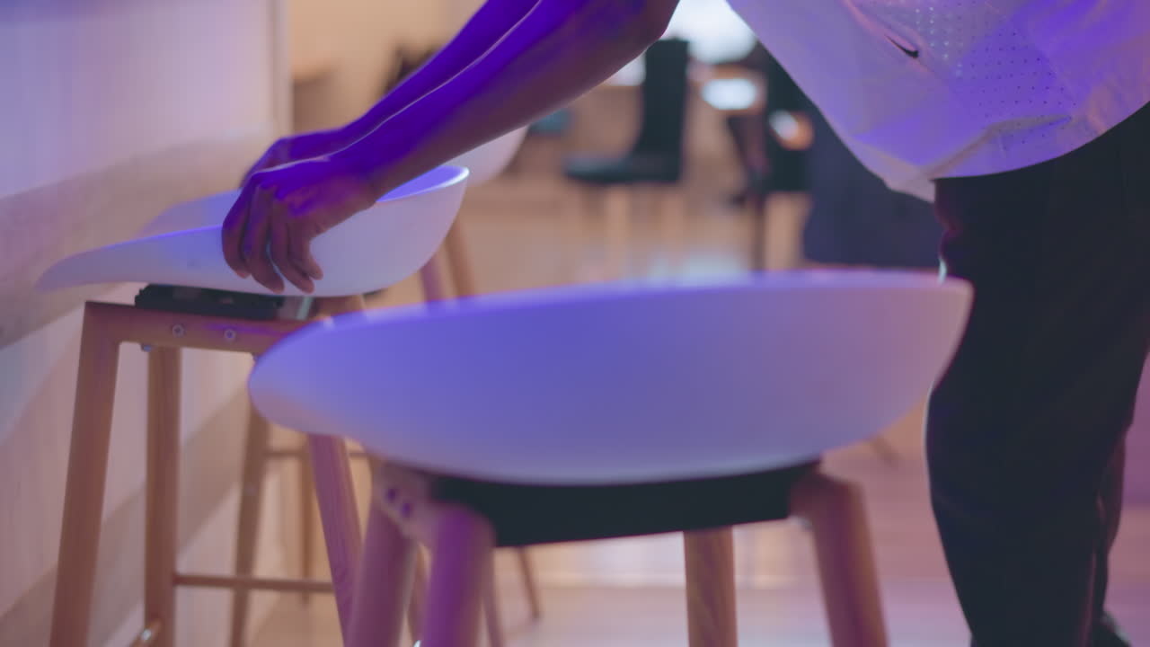 Close-up view of person arranging set of white modern chairs with wooden legs in stylish interior space. Ambient lighting highlights minimalist design with soft purple hues