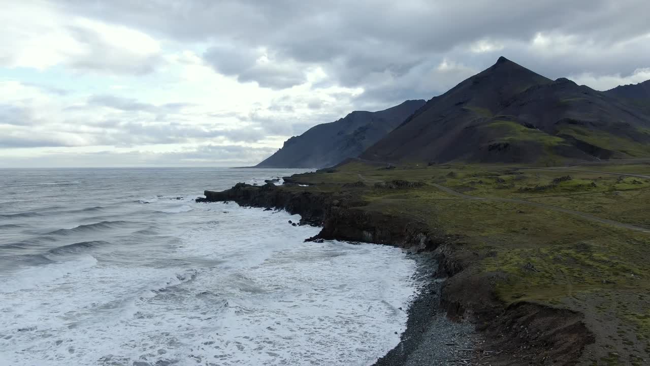 Drone shot of rugged Iceland coastline with waves crashing against rocky shore and steep mountains in background. Cloudy sky and dramatic landscape create moody atmosphere near ocean cliffs