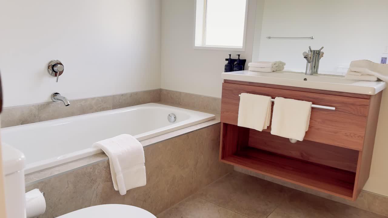 Camera smoothly enters a bright, contemporary bathroom with a bathtub, wooden vanity, towels, and mirror. Natural daylight illuminates the clean, minimalist space
