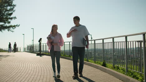 students walking on interlocked path near railing, holding notebooks, engaging in cheerful conversation with blurred group of people in background under bright sunlight