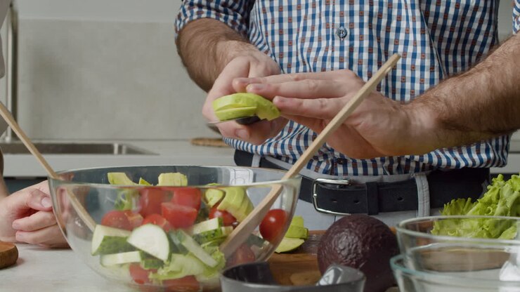Close Up Of Man Hands Adding Avocado Slices In A Glass Bowl To Prepare An Healthy And Tasty Salad