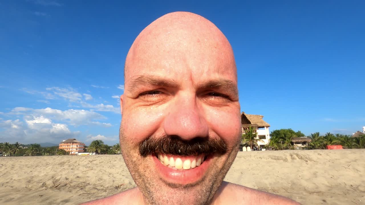 primer plano de un hombre caucásico con bigote sonriendo frente a la cámara en la playa
