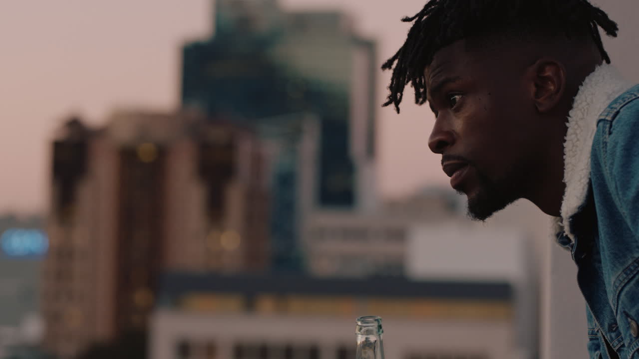 giovane afroamericano attraente che beve alcol godendosi la vista sul tetto dello skyline della città guardando contemplativo al tramonto