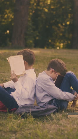 funny junior schoolboys with textbooks and schoolbags sit on lush lawn resting after preparing for exam in park
