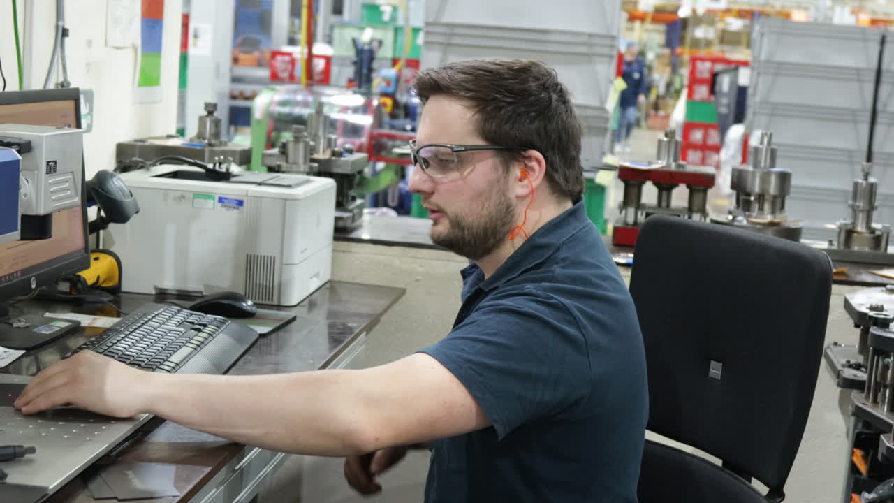 hombre escribiendo en una computadora en un almacén industrial, maquinaria en el fondo, día