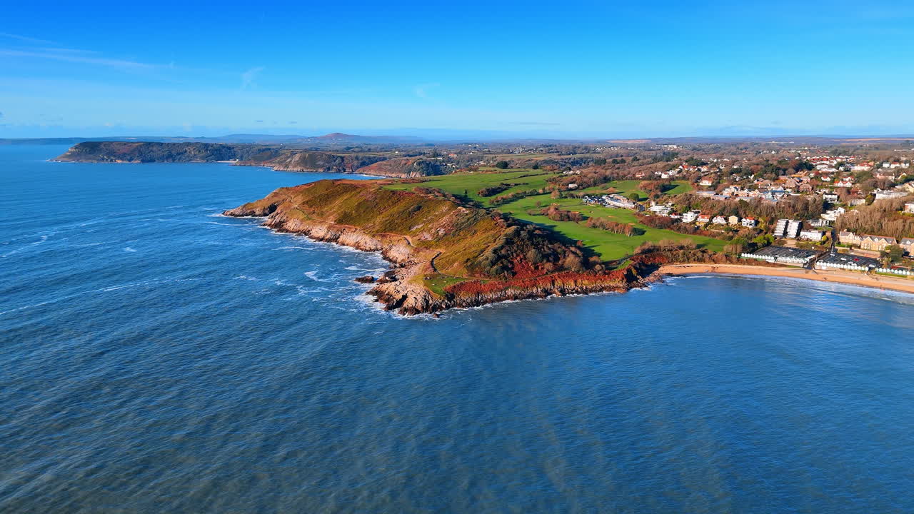 Toma aérea ampla com drone da costa e do promontório da Baía de Caswell. Vista aérea da costa galesa acidentada, com falésias verdes, uma baía de areia e áreas residenciais distantes.