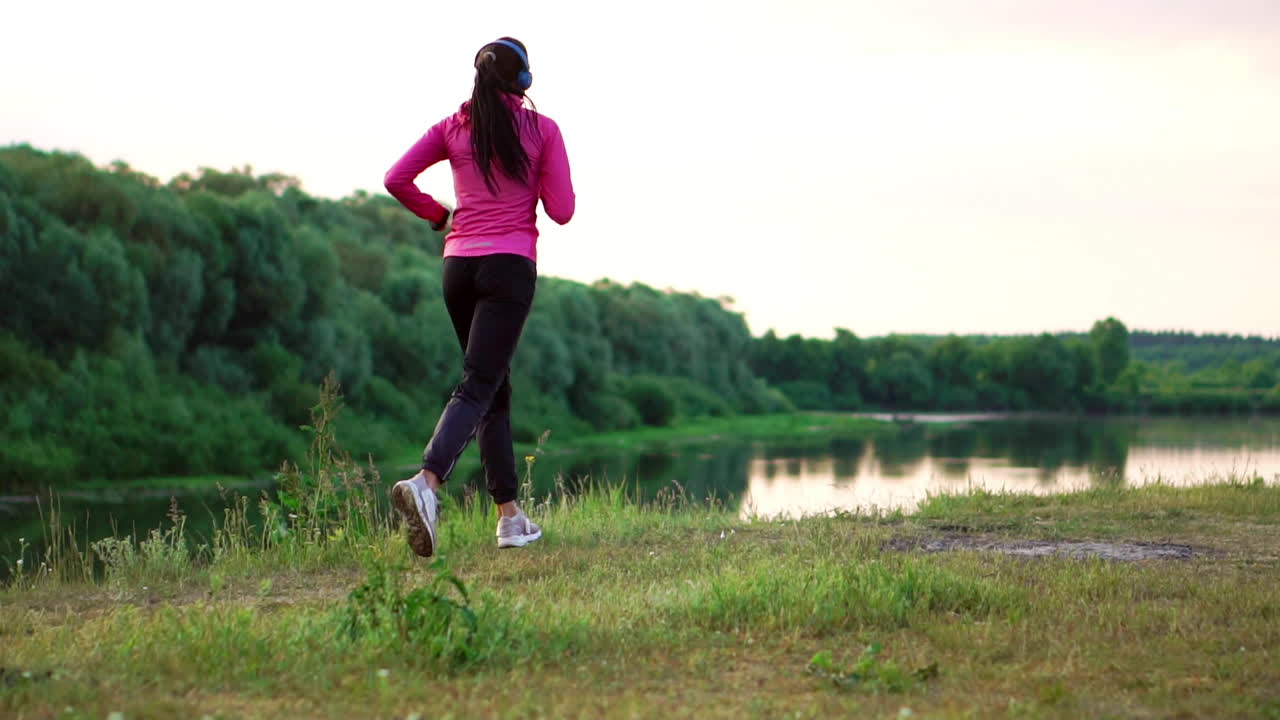 Una ragazza con una giacca rosa e pantaloni neri corre vicino al fiume, con le cuffie, preparandosi per la maratona.