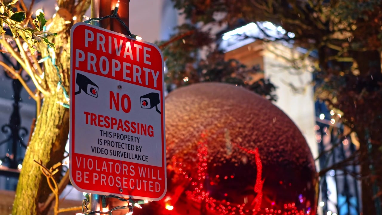 Private property warning sign with Christmas decorations in background night. Red metal sign with security camera icons in front of festive holiday lights and a large red bauble at night.