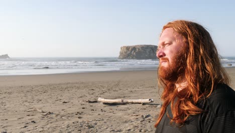 a red-haired man closes his eyes while looking out at the beach enjoying the moment