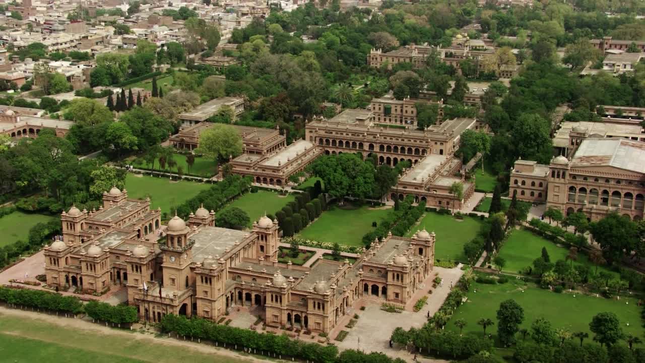 A broad aerial view of a historical university campus in Pakistan featuring grand colonial architecture surrounded by lush greenery. The campus is set within an expansive cityscape
