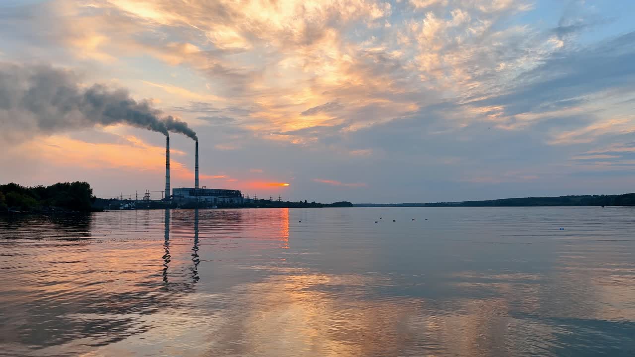 Solnedgang over vand med røg, der stiger op fra et kraftværk nær kystlinjen. Røg stiger op fra skorstene på et kraftværk, mens solen går ned over roligt vand, der reflekterer farverne på himlen