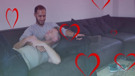 animation of hearts over diverse male couple watching tv