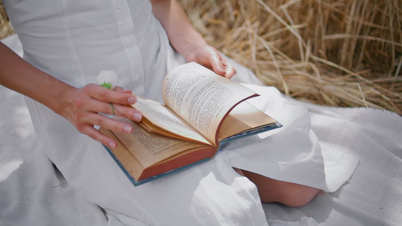 Женские руки переворачивают страницы книги на фоне ржаных колосьев крупным планом. Женщина кладет цветок.