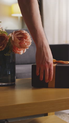 video vertical de un hombre poniendo un regalo en una caja envolta con un regalo decorada con una cinta con una tarjeta de agradecimiento y flores en la mesa en el salón