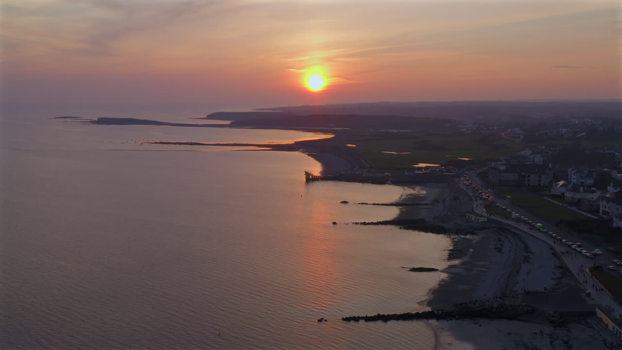 Slow motion aerial approach of waves rolling onto the Salthill shore, Blackrock Diving Board visible in the distance at sunset dusk with orange pink hues