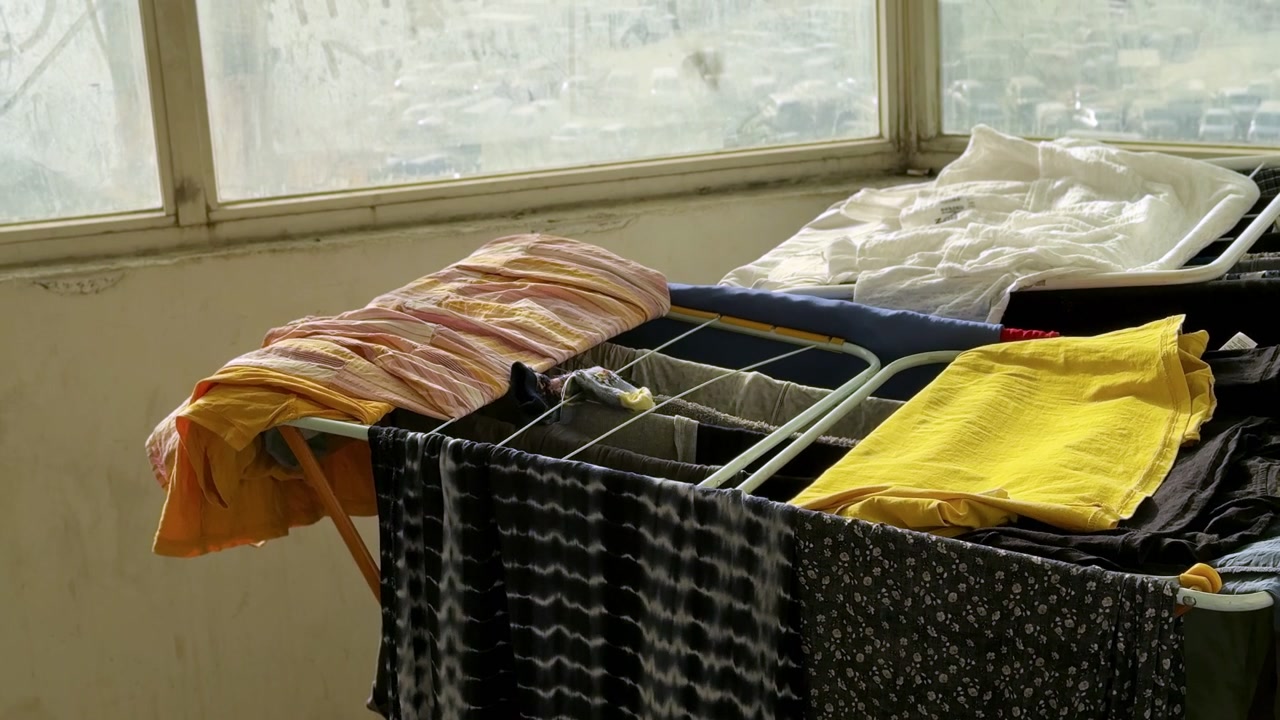 Colorful wet laundry hangs drying on a folding metal rack indoors. Natural daylight illuminates the washed clothes placed next to large dusty windows overlooking a blurred urban street.