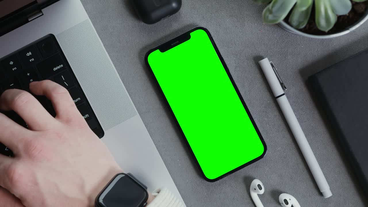 Persona che lavora al laptop con un telefono con schermo verde e vari articoli da ufficio