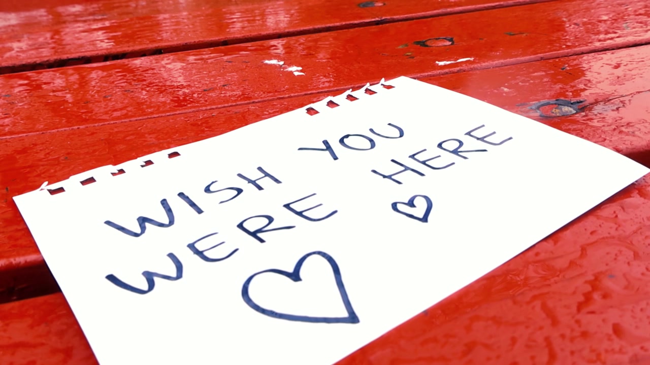 Close up of a handwritten note with the message Wish you were here on a red park table getting wet from the falling rain drops