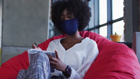 mujer afroamericana con máscara facial sentada en un café escribiendo en un cuaderno