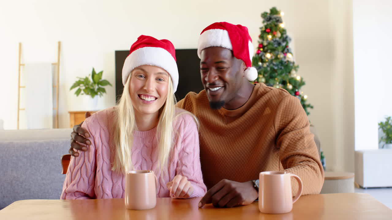 Smiling diverse couple wearing Santa hats enjoying hot drinks by Christmas tree