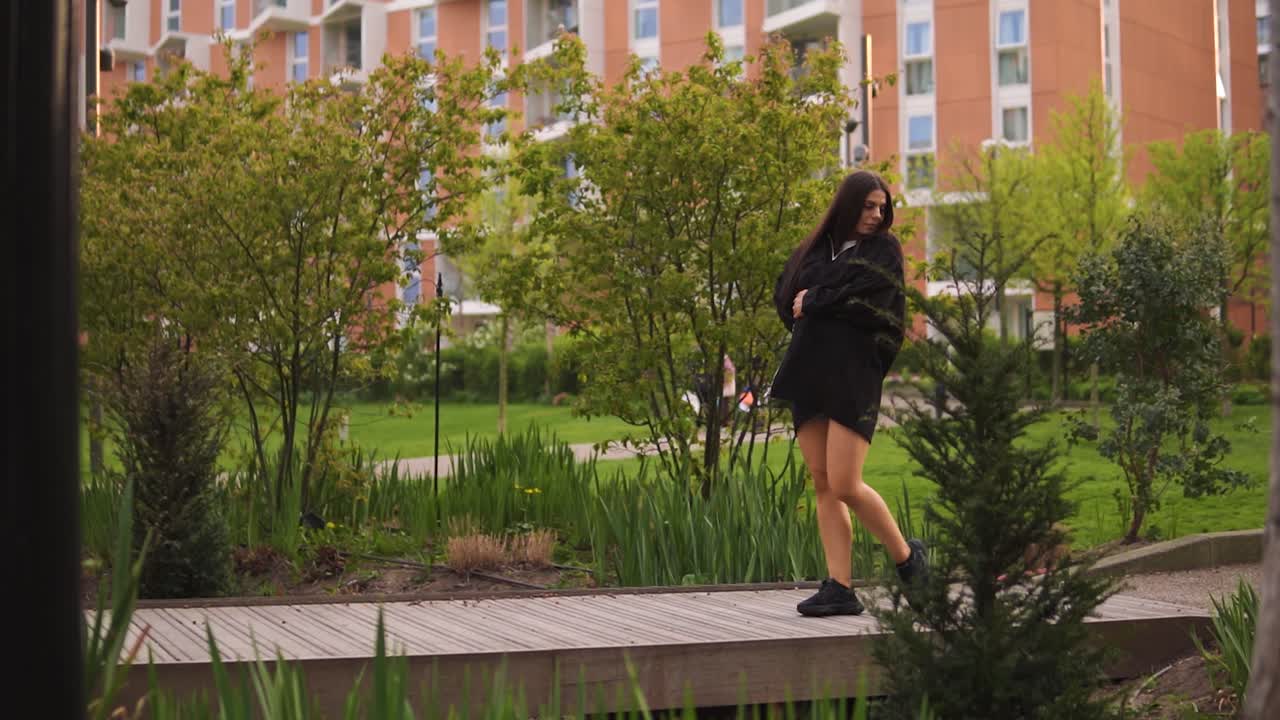 Fashionable female with brown hair is walking across the bridge looking around touching her body wearing jacket and black trainers enjoying time in nature with beautiful building slow motion