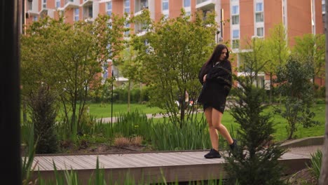 mujer de moda con cabello castaño está caminando por el puente mirando alrededor tocando su cuerpo usando chaqueta y entrenadores negros disfrutando del tiempo en la naturaleza con un hermoso edificio a cámara lenta