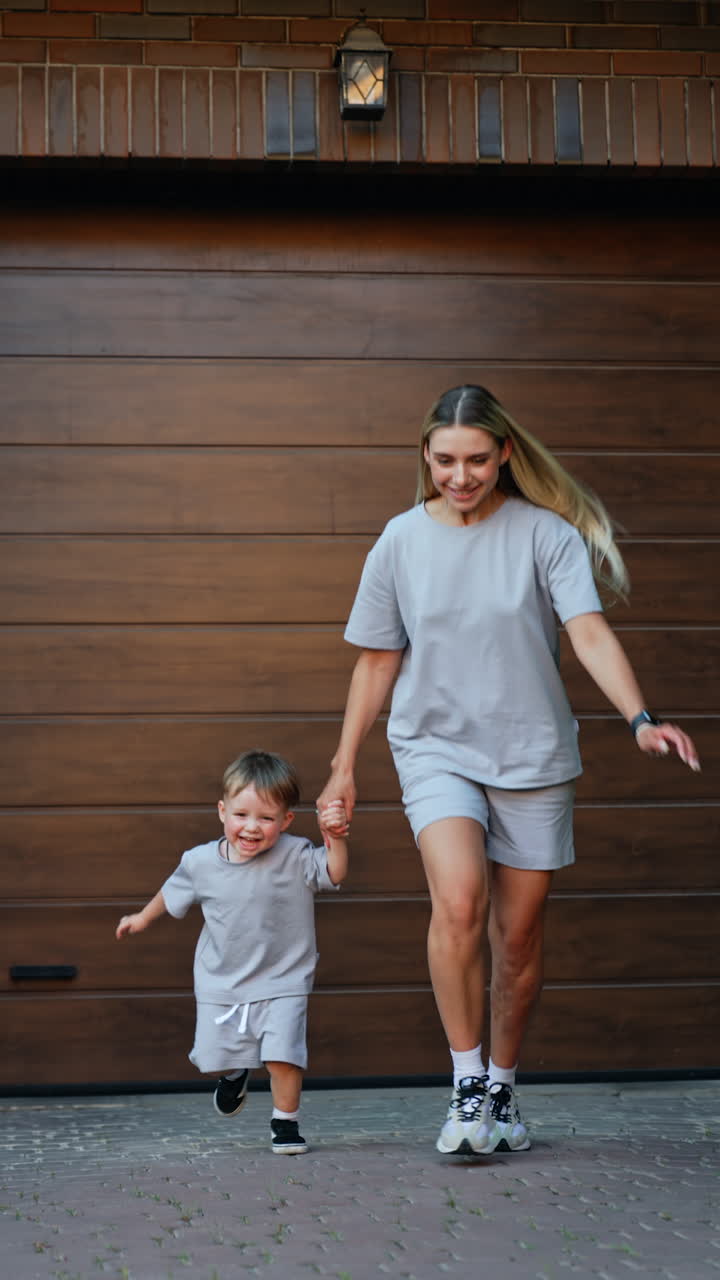 Blonde woman and little boy wearing similar clothes stand at the wooden wall. Mother and kid run up to camera holding hands. Vertical video