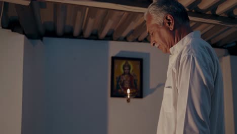 elderly man stands in prayer in a dimly lit room, his gaze directed towards a religious painting illuminated by the warm glow of candlelight, creating a serene and devotional atmosphere