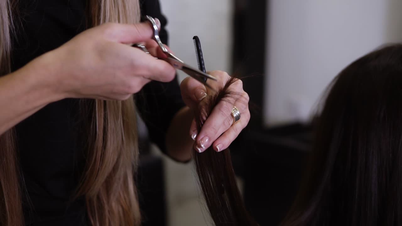 Closeup of skilled hairdresser with french manicure cuts client brown hair ends with new metal scissors using a special technique. Professional stylist taking care of clients ends. Slow motion