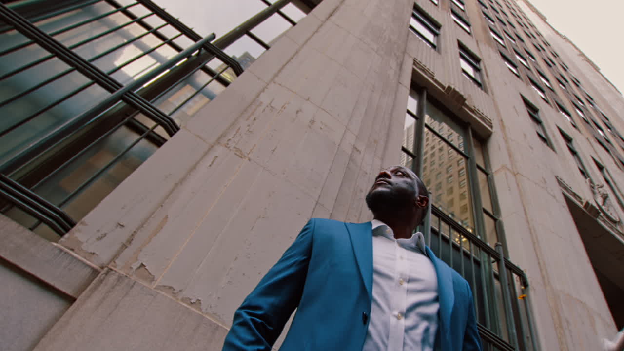 New York, USA, 1 April 2026: Black businessman looking up beside Manhattan office tower. Low angle view of Black businessman in blue suit near stone and glass office buildings in New York