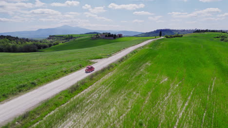 Fast-aerial-following-red-car-on-country-road-by-green-Italian-fields